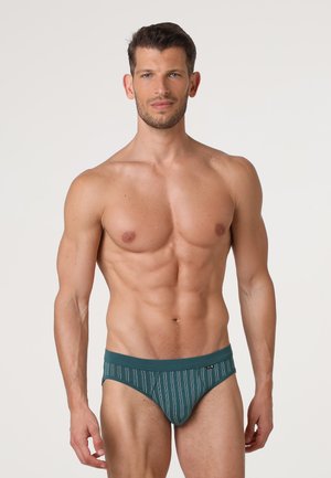 Uomo muscoloso in piedi che indossa slip a righe turchesi e grigie contro uno sfondo chiaro uniforme.