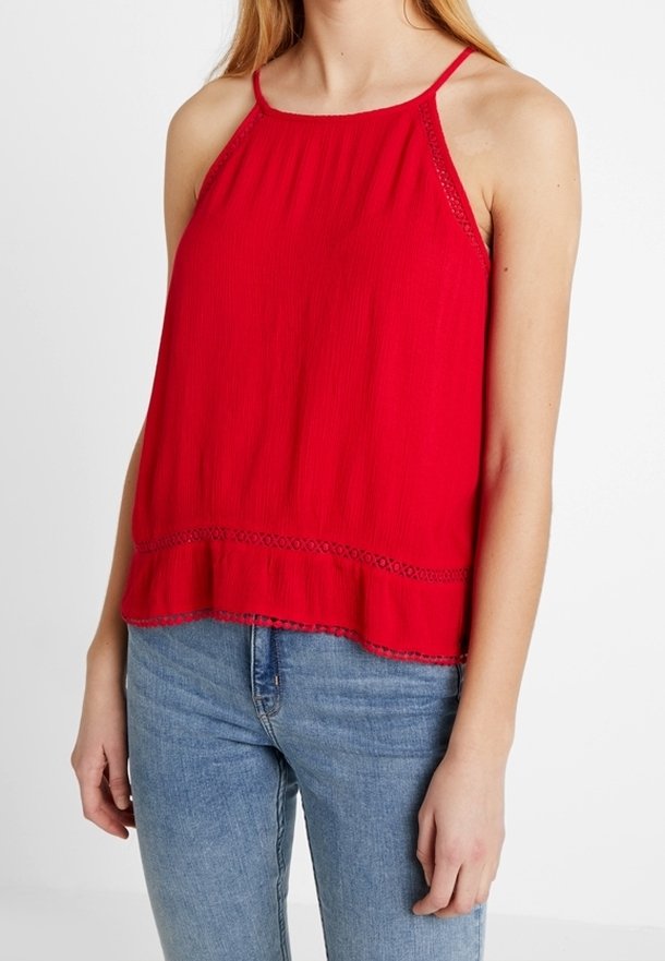 Femme portant un haut rouge sans manches à col licou orné de dentelle, associé à un jean en denim bleu clair, sur un fond blanc.