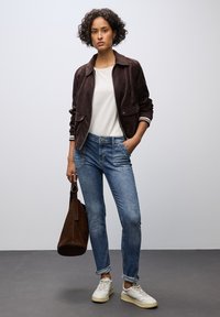 Chaqueta de ante marrón con dos bolsillos frontales, camiseta blanca, vaqueros de mezclilla azul claro y zapatillas blancas. Bolso de hombro marrón en la mano.