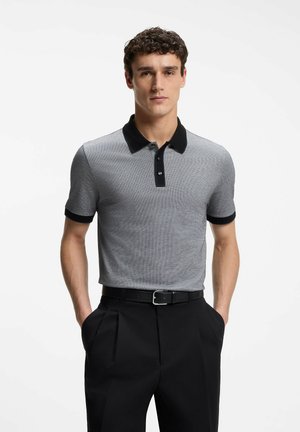 Mann trägt ein graues Kurzarm-Poloshirt mit schwarzem Kragen und Manschetten, schwarzen Gürtel und schwarzen plissierten Hosen, Hände in den Taschen, blickt nach vorne.