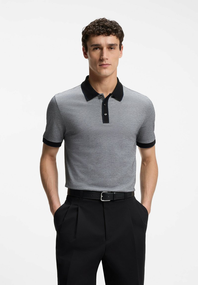 Homme portant un polo gris à manches courtes avec col et poignets noirs, ceinture noire et pantalon noir plissé, mains dans les poches, regardant vers l'avant.
