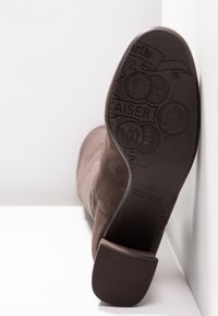 Bottine en daim marron avec un bout légèrement pointu, dotée d'une semelle en caoutchouc texturée et de motifs en relief en forme de pièces de monnaie sous la semelle.