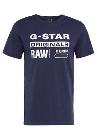 T-paita tummansinisestä puuvillasta, jossa on pyöreä kaula-aukko, lyhyet hihat, ja siinä on suurta valkoista tekstiä: "G-STAR ORIGINALS RAW DENIM" sekä yksinkertainen ääriviivakuva.