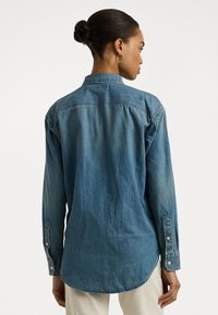 Vidēja zila džinsa krekls ar apkakles kaklu, garām piedurknēm un izliekta apakša. Redzamais šuvums un pogu akcents uz piedurknēm.