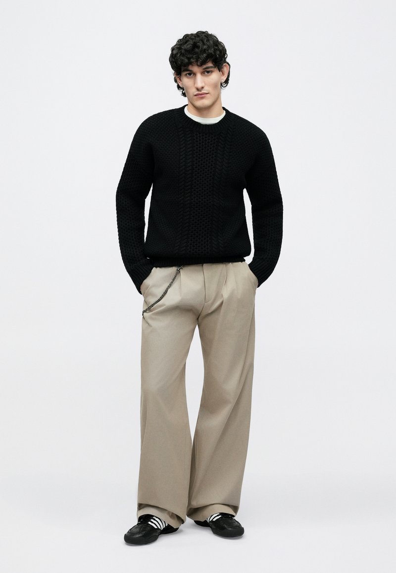 Young man standing with hands in pockets wearing black textured sweater, beige wide-leg trousers with chain, and black sneakers with white stripes.