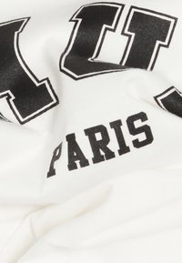 Valkoinen kangas, jossa on mustilla ja suurilla kirjaimilla teksti "PARIS" ja osittain suuria ääriviivakirjaimia tekstuurilla.