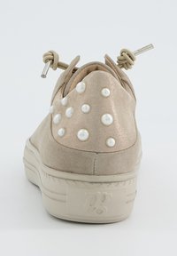 Beige zamšaini sporta apavi ar noapaļu aizmuguri, rotāti ar baltām pērļu detaļām un plakanu gumijas zoli. Iekļauj unikālu aukliņu aizdari.