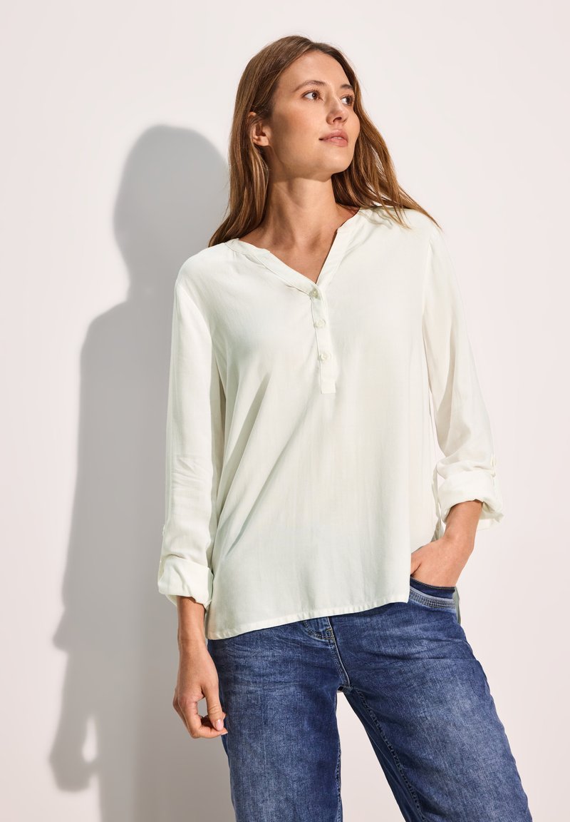 Blusa lunga beige chiaro a maniche lunghe con scollatura a V e bottoni, tessuto morbido, maniche con polsino e vestibilità comoda; abbinata a pantaloni in denim blu.