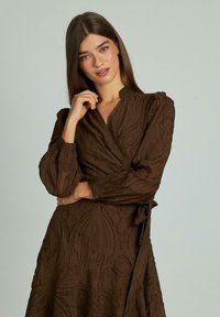 Rochie maro, dintr-un material texturat, cu mâneci lungi și bufante și talie strânsă. Are un model subtil de frunze și o fustă fluidă.