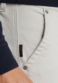 Beige cotton blend pants with a smooth texture, featuring a navy ribbed waistband, metal button, and a black label with "Pale Legend" branding.