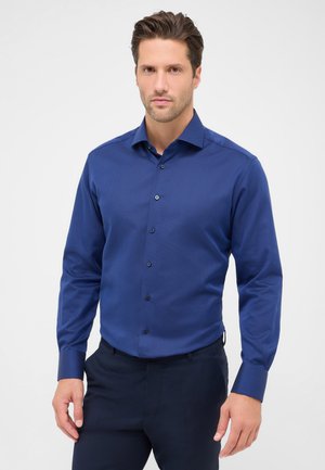 Blaues Langarmhemd aus strukturiertem Stoff mit einem Spread-Kragen, Knopfleiste und dunklen Knöpfen, kombiniert mit dunklen Hosen.