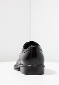 Chaussure habillée en cuir noir à finition lisse, avec un bout arrondi et des accents décoratifs perforés à l'arrière et sur le côté. Talon carré bas.
