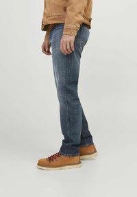 Botas de senderismo marrón claro con cordones rojos, combinadas con jeans ajustados azul oscuro, mostradas de lado contra un fondo sencillo.