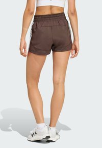Shorts de sport marron avec une ceinture élastique, des bandes latérales et une texture lisse, associés à des chaussures de sport blanches et noires.