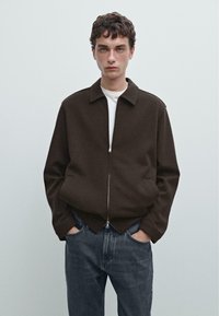 Brązowa kurtka na zamek z kołnierzem, noszona na białym T-shircie. Model ma ręce w kieszeniach, w zestawieniu z ciemnoniebieskimi dżinsami.
