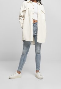 Chemise cream oversize avec bouton et détails de poche, associée à un jean skinny bleu clair et des baskets blanches. Fini en tissu lisse.