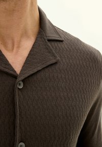 Brown textured shirt with a collared neck, featuring a subtle geometric pattern and button closure. Made of soft, woven fabric.