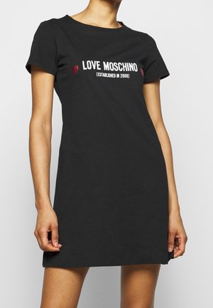 Kvinde iført en sort kortærmet kjole med teksten "LOVE MOSCHINO (ESTABLISHED IN 2008)" og to små røde hjerter på brystet.
