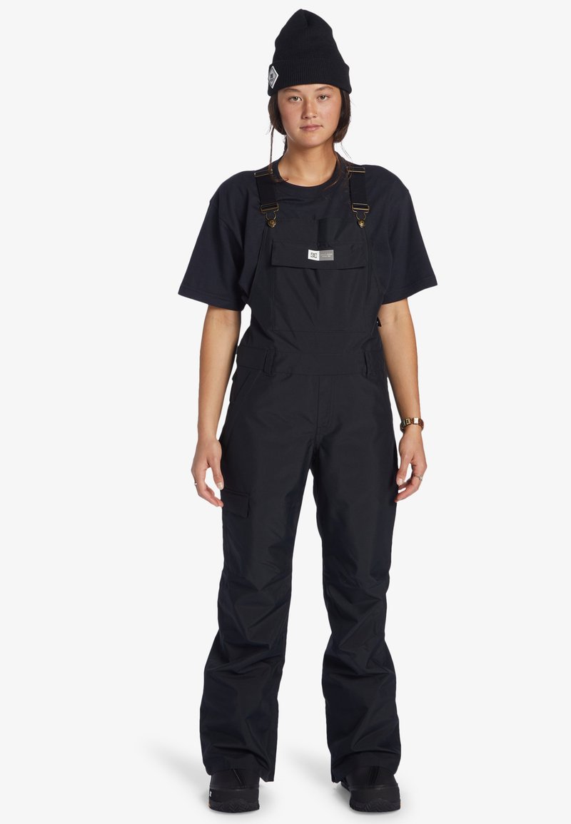 Nuori henkilö, jolla on musta pipo, löysä musta lyhythihainen paita, mustat haalarit ja mustat saappaat, seisoo valkoista taustaa vasten.