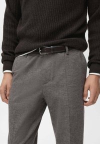 Pantaloni grigi a motivi houndstooth, vestibilità su misura, con pieghe frontali e una cintura in pelle marrone scuro. Maglione scuro a coste indossato sopra.