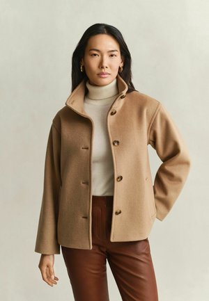 Frau mit langen dunklen Haaren, die goldene Creolen, einen beigen Rollkragenpullover, einen braunfarbenen Mantel mit Knöpfen und braune Hosen trägt.