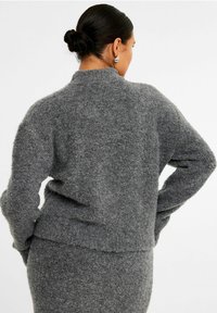 Grå, fuzzy strikket sweater med høj krave og lange ærmer. Har en afslappet pasform og en kant, der ender nær taljen.