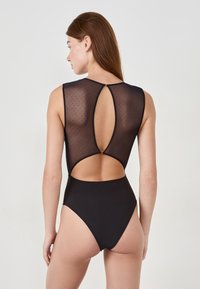 Czarny strój kąpielowy z przezroczystym plecami w kropki, wysokimi wycięciami na nogi oraz dekoltem w kształcie łezki z małym złotym akcentem przy szyi.