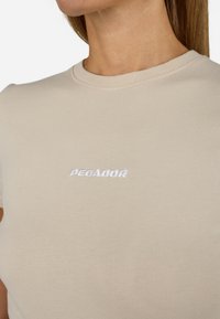 Bež pamukasta majica s okruglim izrezom i kratkim rukavima. Na prednjoj strani sadrži izvezeni natpis "PEGADOR" u beloj boji.
