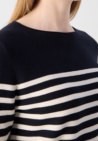 Zbliżenie osoby w czarno-białym swetrze robionym na drutach w poziome paski, z okrągłym dekoltem, ukazujące obszar szyi i ramion.