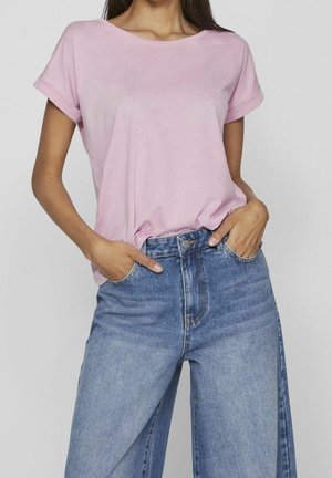 Femme portant un t-shirt à manches courtes rose clair, rentré dans un jean bleu taille haute à jambes larges, les mains dans les poches.