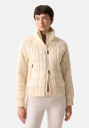 Frau trägt eine hellbeige gesteppte Jacke mit gerippten Bündchen und Kragen, kombiniert mit passenden beigen Hosen, steht vor einem schlichten Hintergrund.