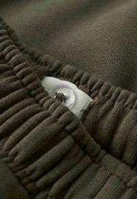 Olīvzaļa auduma ar ribotu tekstūru, ar elastīgu jostas daļu un baltu birku ar caurspīdīgu pogu detaļu.