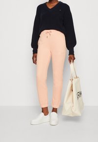 Persoon in een marineblauwe trui, lichtroze joggingbroek, witte instappers, met een crèmekleurige tas met zwarte tekst, tegen een effen achtergrond.