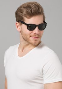 Černé sluneční brýle s tmavými skly. Plastový rám s matným povrchem. Bílé tričko s výstřihem do V. Neutrální šedé pozadí.