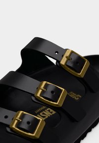 Μαύρο δερμάτινο σανδάλι με τρεις ρυθμιζόμενους ιμάντες, που διαθέτει αγκράφες από μέταλλο σε χρυσαφί χρώμα και υφή στη μαύρη σόλα.