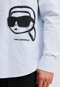 Striped blue and white button-up shirt featuring a black graphic of a person with sunglasses and a name "KARL LAGERFELD" printed below.