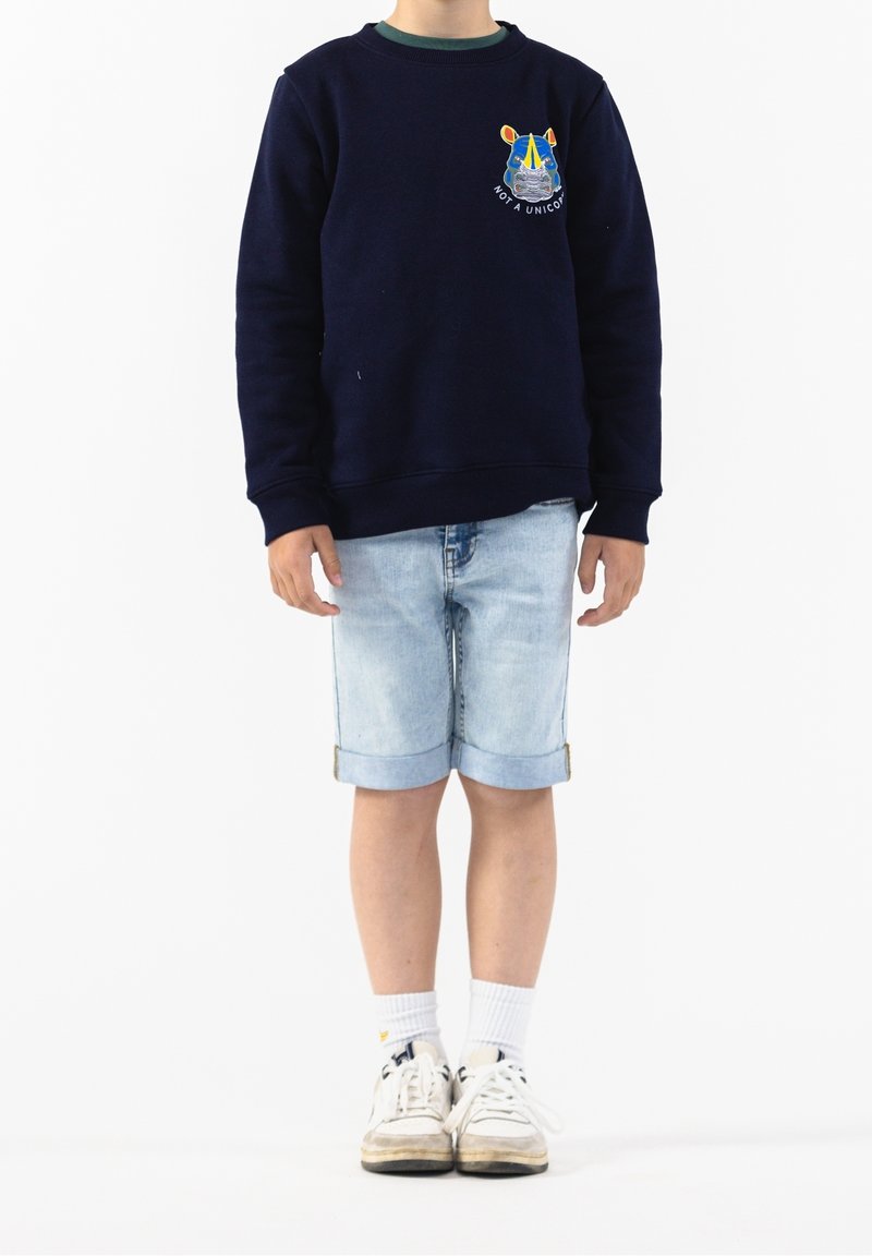 Sweat-shirt bleu marine avec motif brodé, col rond ; short en denim clair avec ourlets retroussés ; baskets blanches avec accents gris.