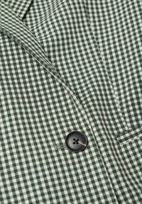 Tela de cuadros vichy en verde y blanco, con un botón negro de cuatro agujeros, doblada mostrando una ligera textura y líneas definidas.