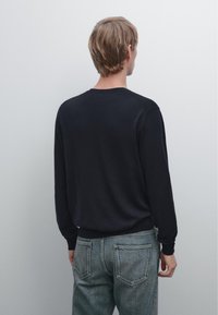 Persona vista di spalle, che indossa una camicia nera a maniche lunghe e jeans blu sbiaditi, in piedi contro uno sfondo grigio chiaro uniforme.