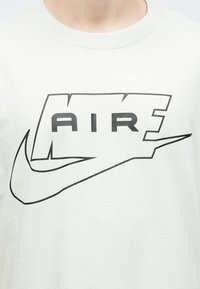 Tricou alb cu text mare "NIKE AIR" conturat în negru și logo swoosh centrat pe piept, purtat de o persoană.