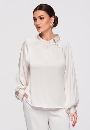 Kobieta o ciemnych włosach związanych w niski koczek, ubrana w luźną białą bluzkę z ozdobnymi guzikami oraz dopasowane białe spodnie.