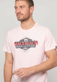 T-shirt rose clair à manches courtes avec un graphique au style vintage "Maui Beach" en rouge et noir. Tissu en coton doux, design à encolure ras du cou.