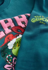 Pull vert avec Le Grinch portant un chapeau de Père Noël, un sucre d'orge, et le texte "The Grinch Dr. Seuss" en jaune et vert.