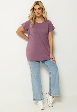 Lyshåret kvinde står op ad en væg iført en løs lilla t-shirt, lyseblå vidde jeans og hvide sneakers.