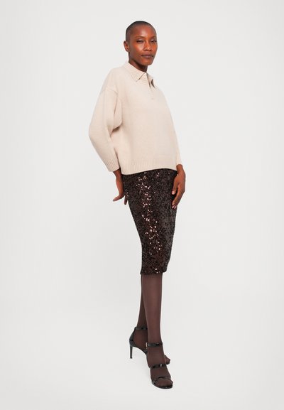 Woman wearing a beige sweater, black sequin pencil skirt, black tights, and black high-heeled sandals, standing against a white background.