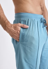 Helle blaue Loungehosen mit einem elastischen Bund, Kordelzugverschluss, Seitentasche und Markenetikett an der linken Naht. Weiche Textur.