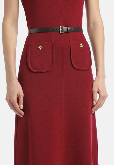 Rdeča pletena obleka z oprijetim vrhom in razširjenim dnom, ima dva velika sprednja žepa z zlatimi gumbi in rjavi usnjeni pas v pasu.