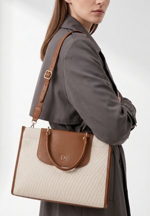Femme en trench-coat gris portant un grand sac cabas en cuir beige et marron avec bandoulière et poignées supérieures.