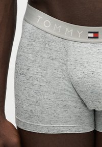 Gros plan sur un boxer gris de la marque Tommy porté par une personne à la peau foncée, montrant la ceinture avec le nom et le logo de la marque.