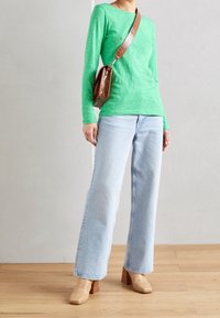 Maglietta verde a maniche lunghe, jeans a gamba larga blu chiaro e scarpe con tacco color cammello con una texture a coccodrillo. Borsa a tracolla marrone con una tracolla intrecciata.